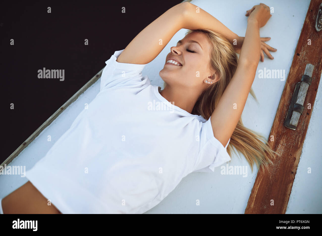 Attractive young woman in a bikini bottom and shirt lying with her eyes closed on the deck of a yacht catching a suntan Stock Photo
