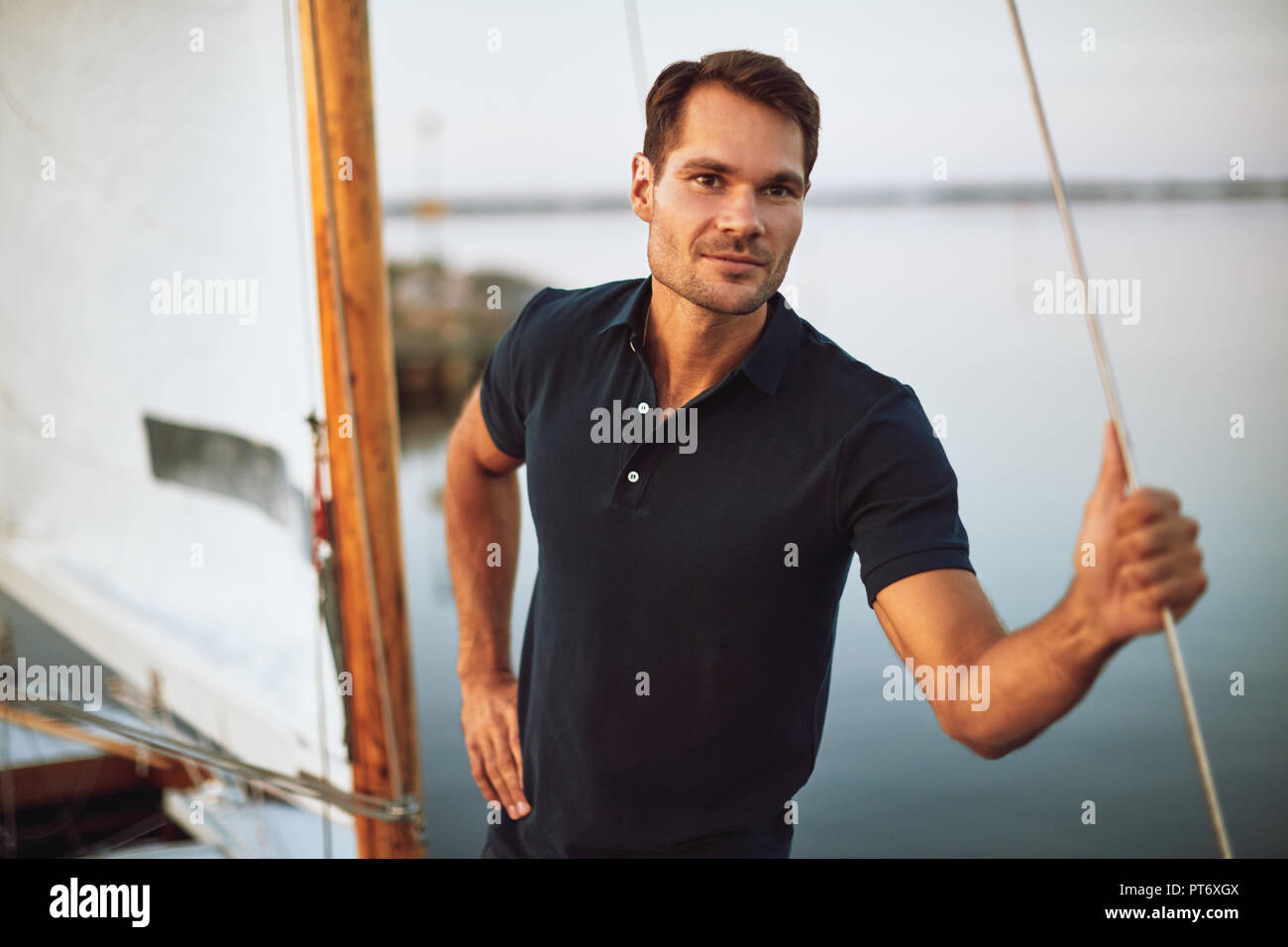 Handsome young man leaning on some rigging while standing alone on the deck of his yacht enjoying a sunny day sailing Stock Photo
