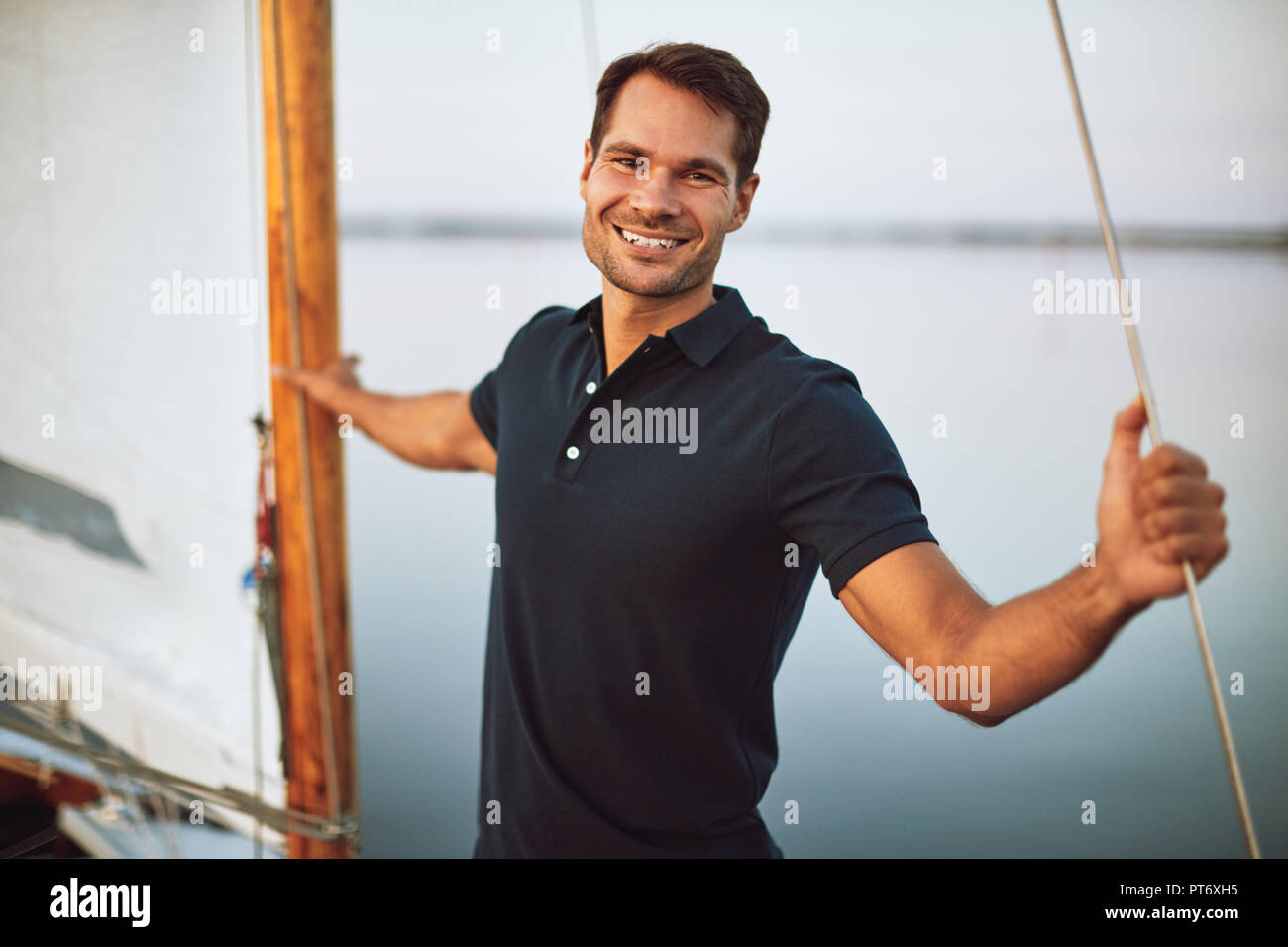 Smiling young man leaning on some rigging while standing alone on the deck of his yacht enjoying a sunny day sailing Stock Photo