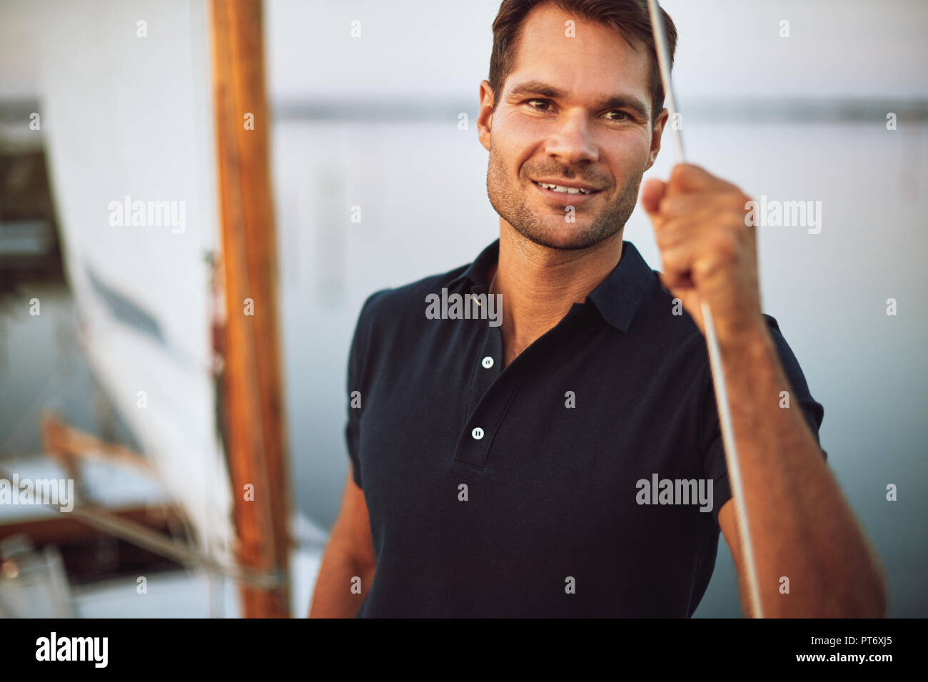 Handsome young man leaning on the rigging of his yacht and smiling while out for a sail Stock Photo