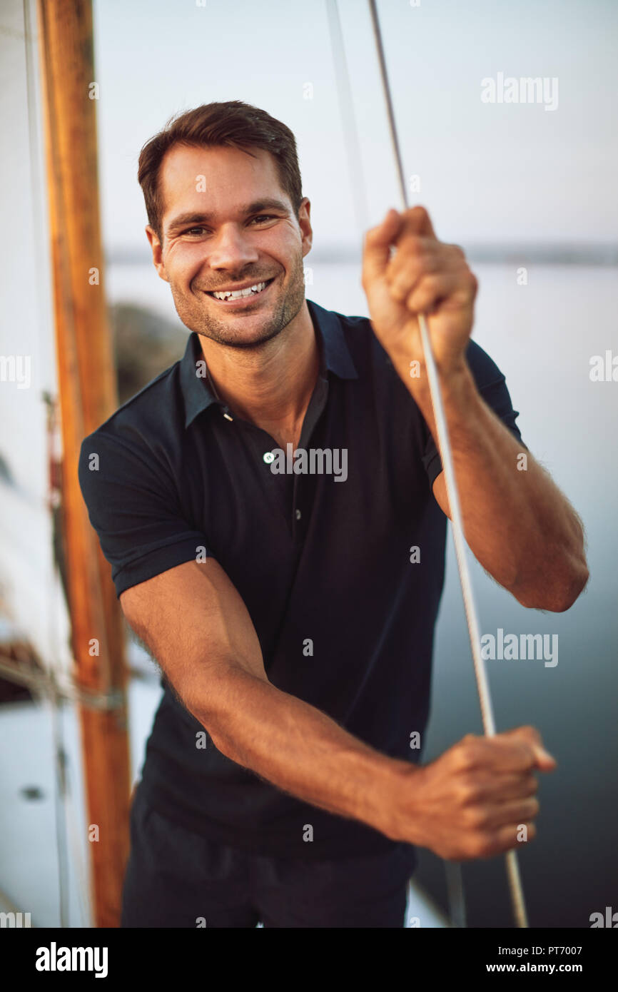 Smiling young man leaning on some rigging while standing on the deck of his yacht enjoying a sunny day sailing Stock Photo