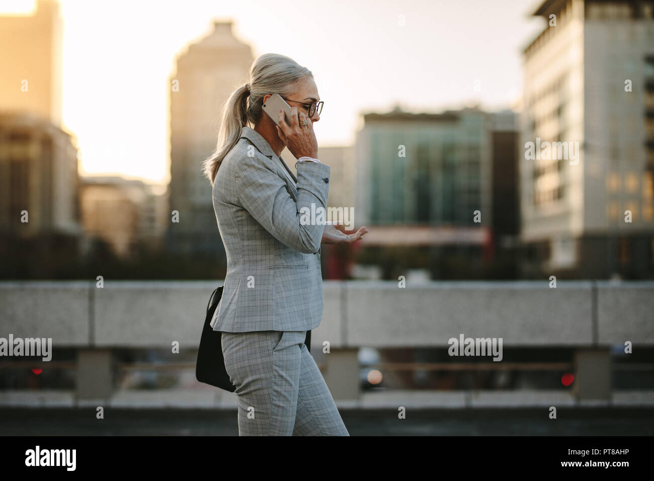 Side view of mature woman in business suit walking back to home talking on phone. Senior businesswoman using cellphone on city street with urban backg Stock Photo