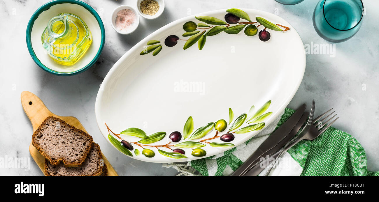 banner of Empty handmade oval dish with olive tree branches on a white marble table. olive oil and cutlery. Mediterranean table setting Stock Photo