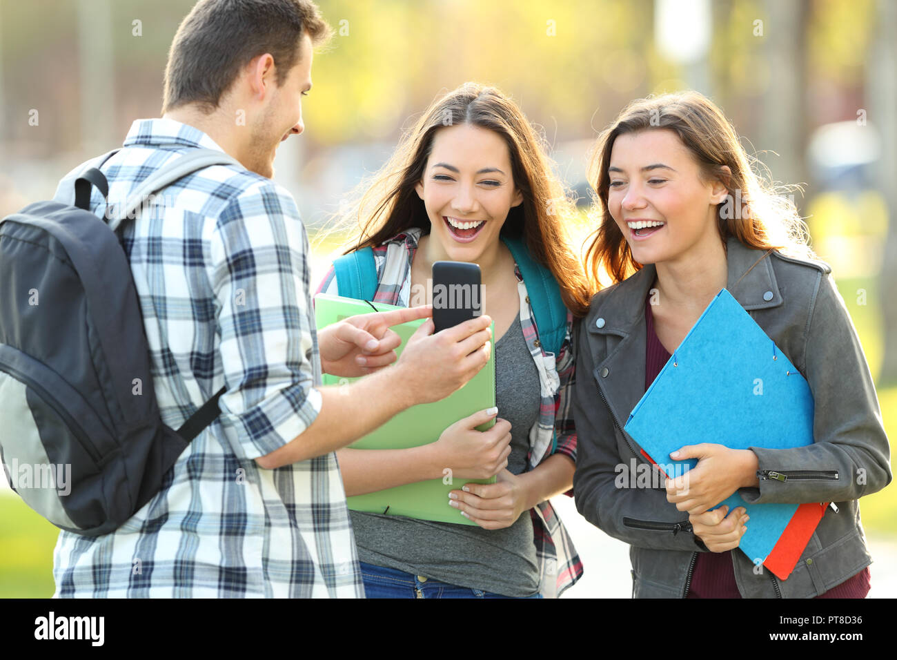 Three happy students sharing smart phone content in a park Stock Photo