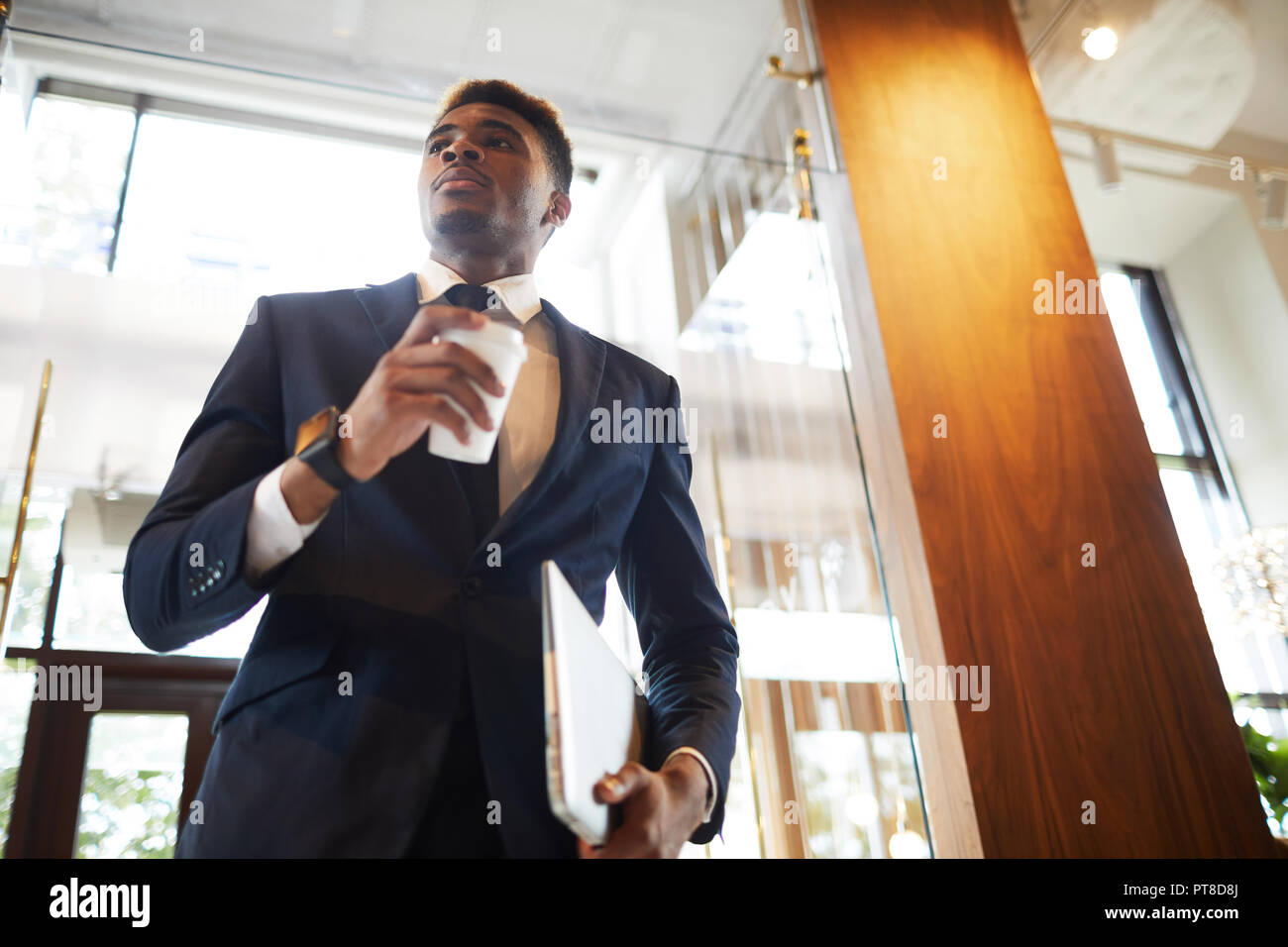Below view of serious confident modern businessman in formal suit holding laptop and drinking coffee from disposable cup while standing in lobby Stock Photo