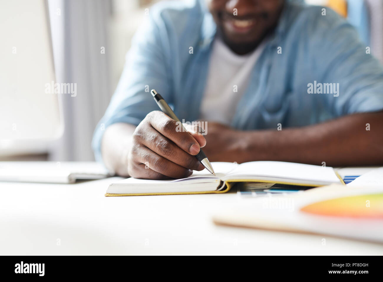 African-american creative guy with pen making working notes in notebook while sitting by desk Stock Photo