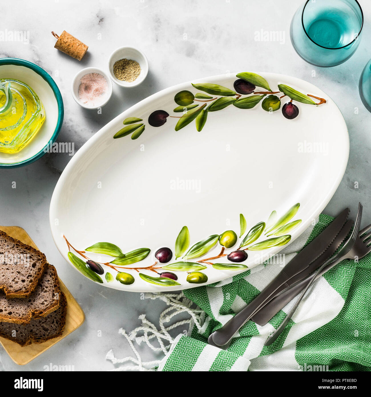 Empty handmade oval dish with olive tree branches on a white marble table. olive oil and cutlery. Mediterranean table setting Stock Photo
