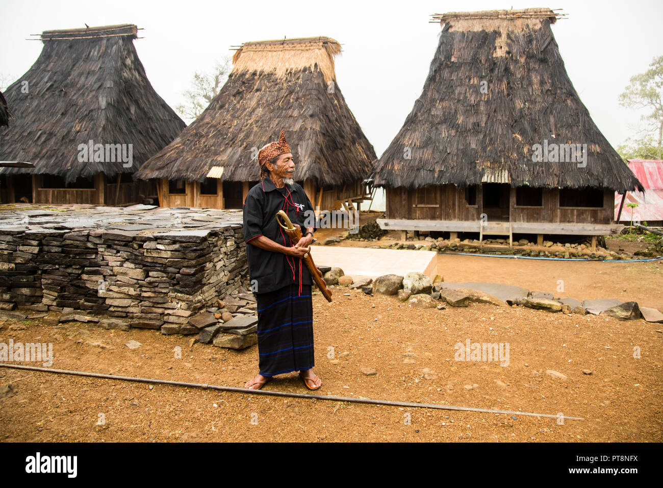 A man in a traditional sarong and red-patterned headscarf holds a wooden object in both hands, standing beside a low stone wall in a mountain village near Moni, Flores, Indonesia. Stock Photo