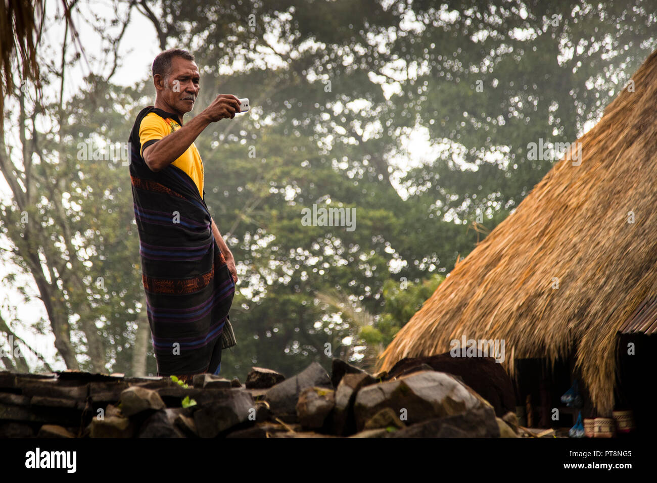 A man in a yellow polo shirt over his ikat sarong takes a photo with his smartphone against dense foliage and a thatched roof in a mountain village near Moni, Flores, Indonesia. Stock Photo