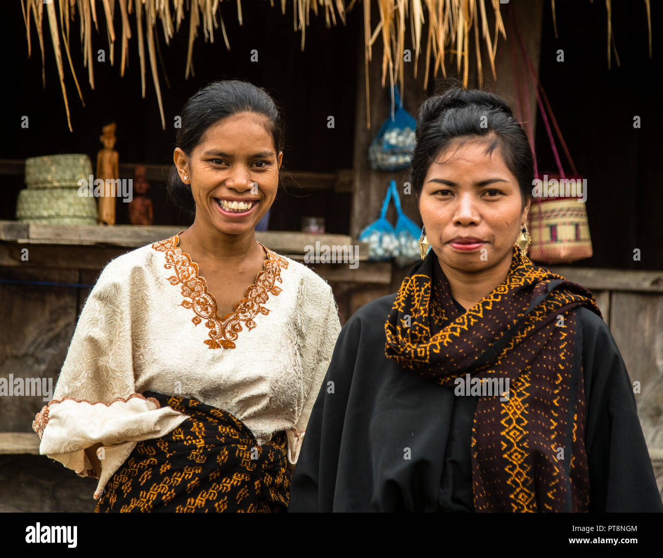 One of the two women smiles broadly while her companion stands beside her with a serious expression, both in traditional ikat clothing in front of a wooden house in a mountain village near Moni, Flores, Indonesia. Stock Photo