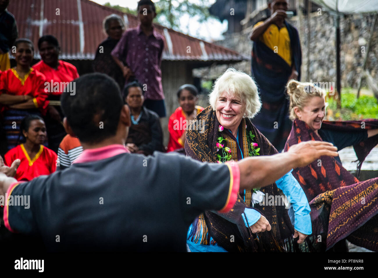 Two visitors join the welcome ritual in Wologai, Flores, Indonesia, alongside the villagers, one of them wearing a flower garland and a dark ikat shawl. Traditional wooden houses of the village stand in the background. Stock Photo