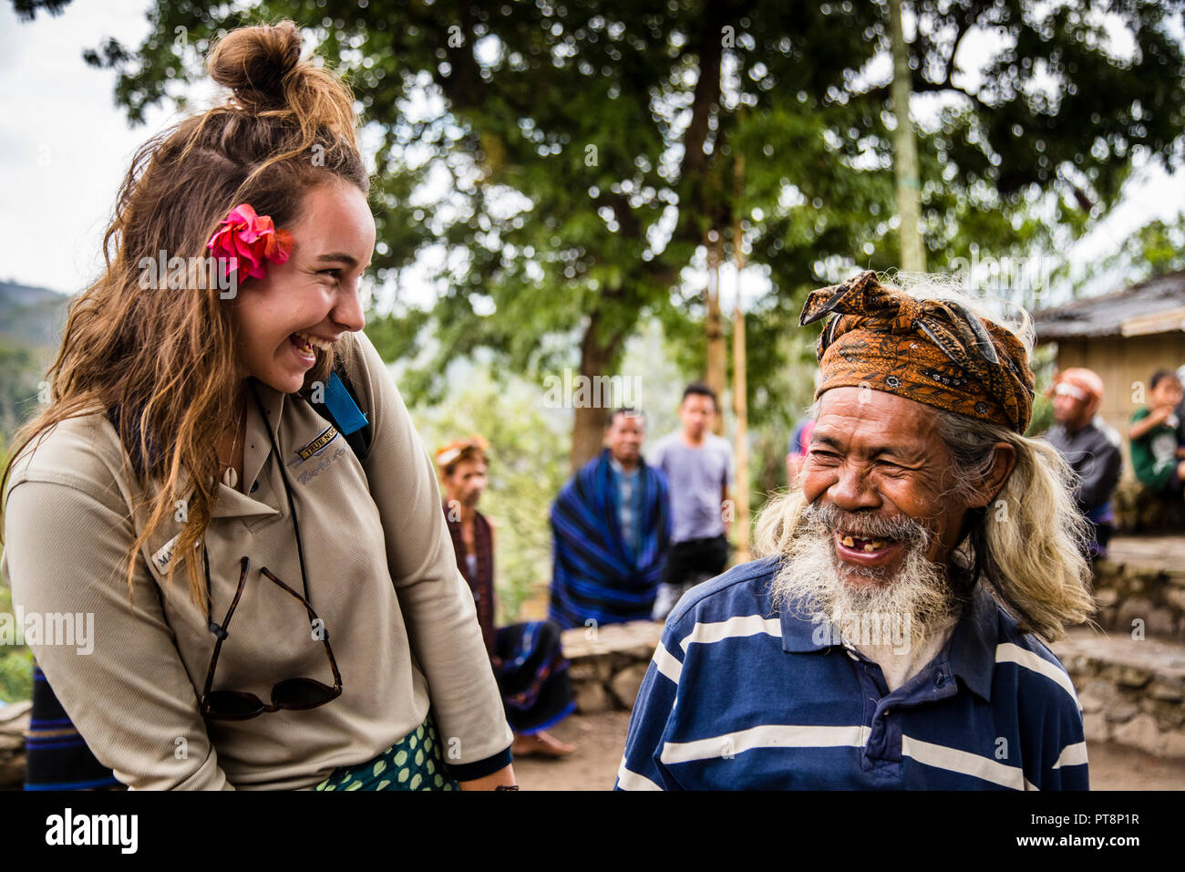A female visitor and an elderly man with gaps in his teeth are laughing together in the village of Wologai on Flores, Indonesia; in the background, villagers are sitting in front of a lumbung house. Stock Photo