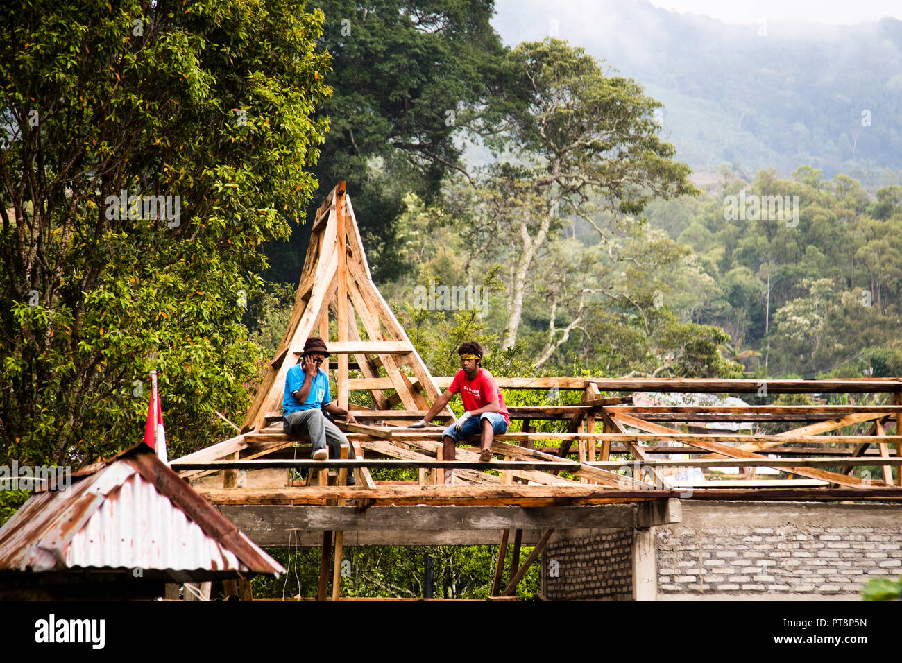 Two carpenters sit on the wooden roof frame of a lumbung storehouse under construction near Ende, Flores, Indonesia, an Indonesian flag flying at the gable. Stock Photo