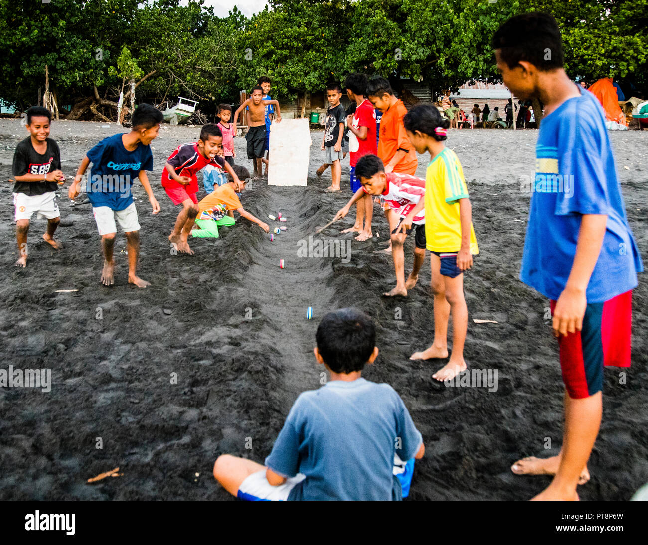 Boys gather around a sand trench with a wooden block and thrown objects during the traditional throwing game on the black-sand beach of Ende, Flores, Indonesia, mangrove trees lining the beach in the background. Stock Photo