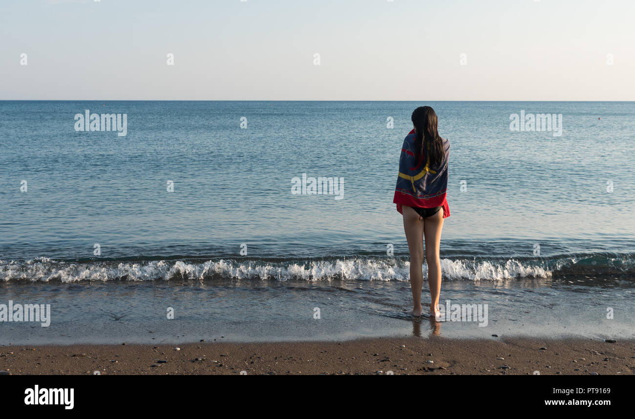 Teen standing at water's edge with towel looking out to sea Stock Photo