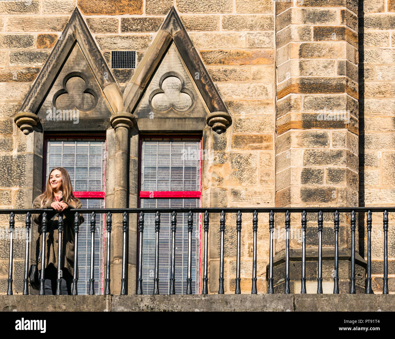 Woman smiling in sunshine standing by railings, in front of old church windows, West Bow, Victoria Street, Edinburgh, Scotland, UK Stock Photo