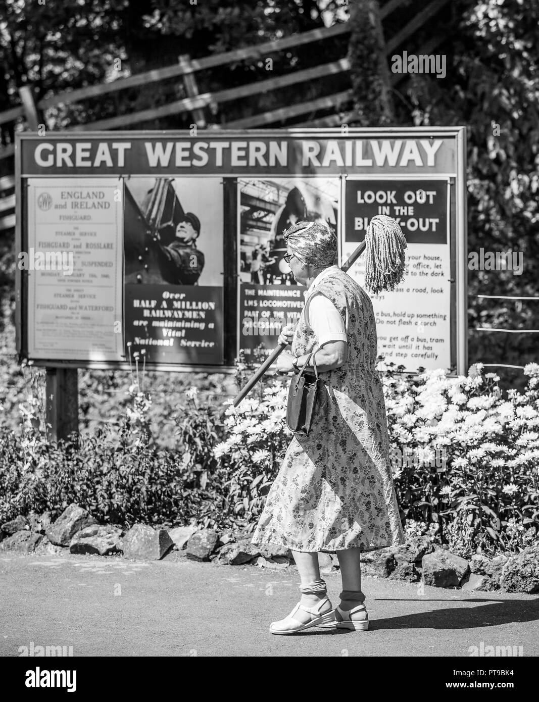 Monochrome rear of woman in overalls, headscarf, wrinkled stockings as vintage cleaning lady with mop at heritage railway station, UK 1940s WWII event. Stock Photo