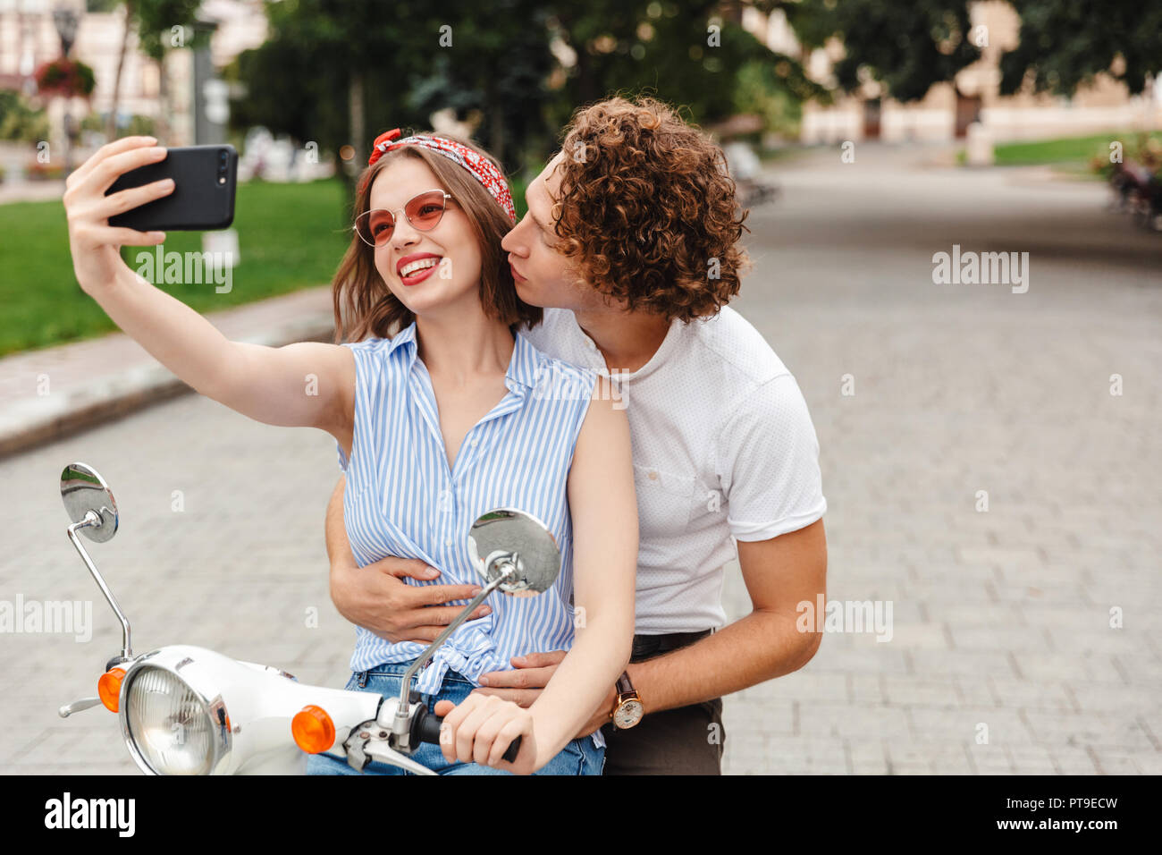 Portrait of a happy young couple riding on a motorbike together at the city street, taking a selfie, kissing Stock Photo
