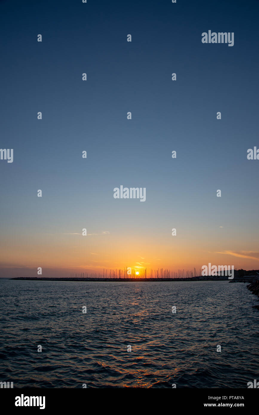 Vertical photo with sunset in Cecina Italy. The sun is behind stone breakwater. Many masts are visible on horizont. Sky is clear with orange and blue  Stock Photo