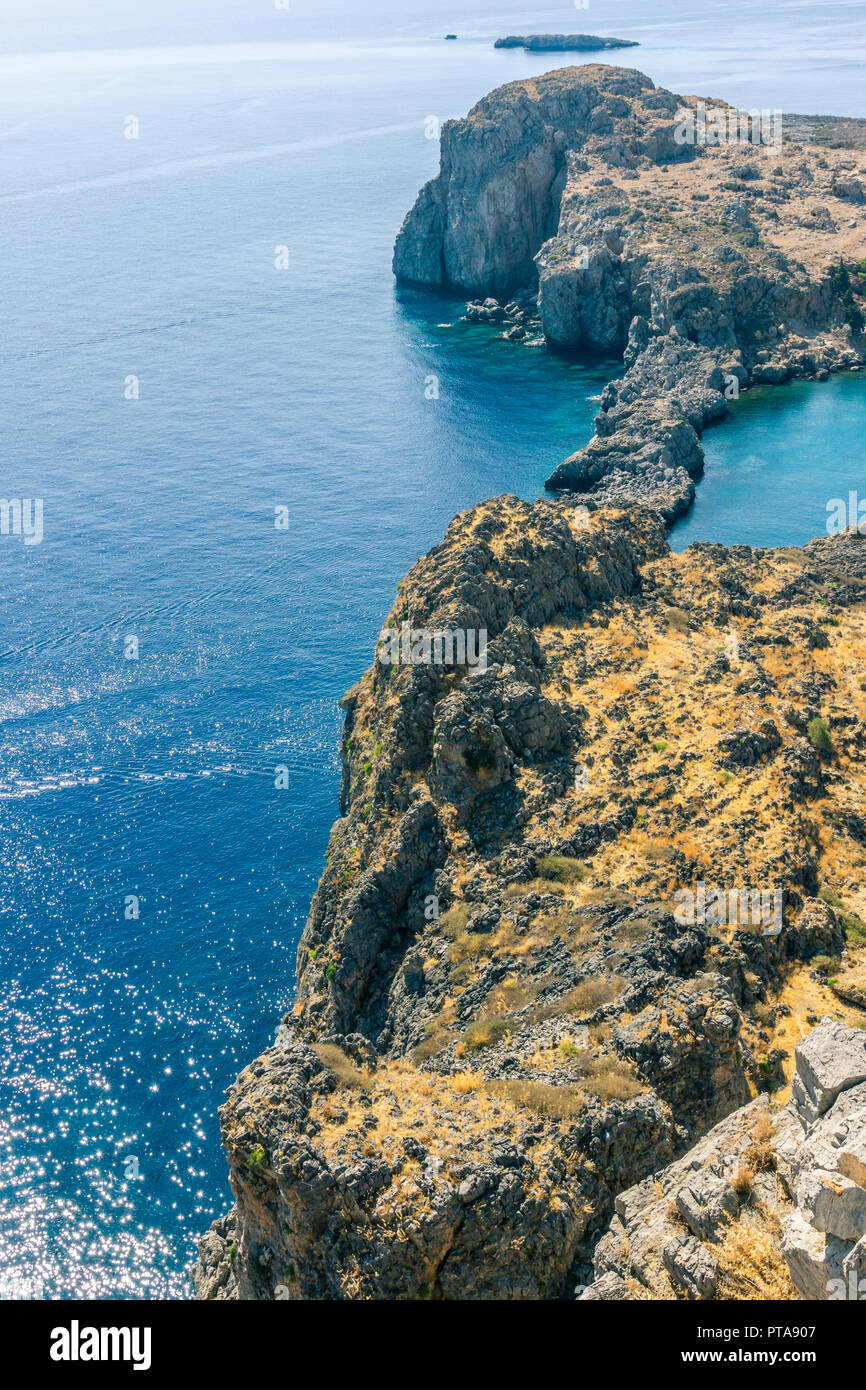 Vertical photo with view down on rocky reef covered by dry grass and with blue sea around. Reef is under acropolis in Rhodos town Lindos captured in s Stock Photo