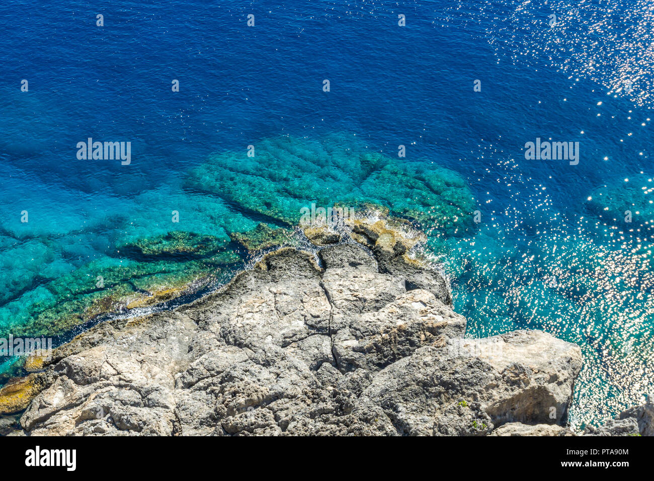 Horizontal photo with view down on rocky reef with blue sea around. Reef is under acropolis in Rhodos town Lindos captured in sunny summer day. Stock Photo