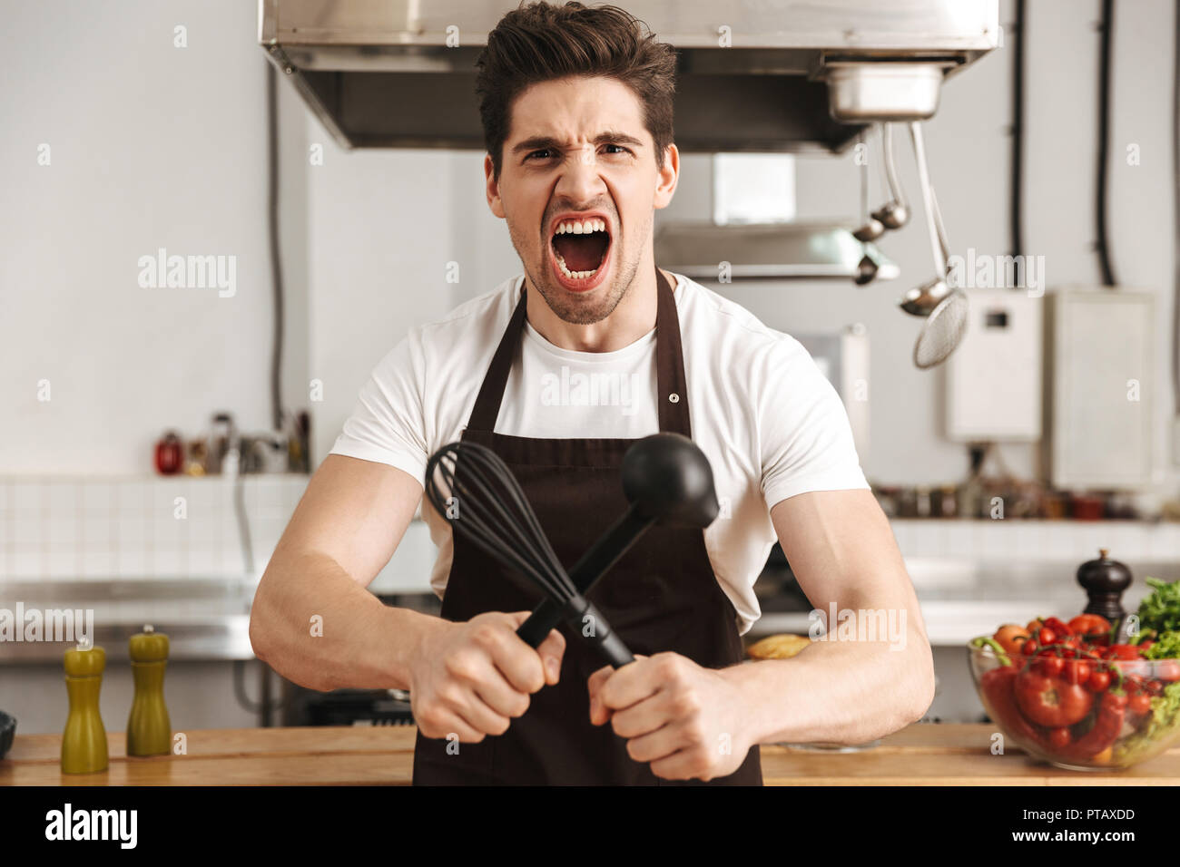Angry young man chef cook in apron standing at the kitchen, holding