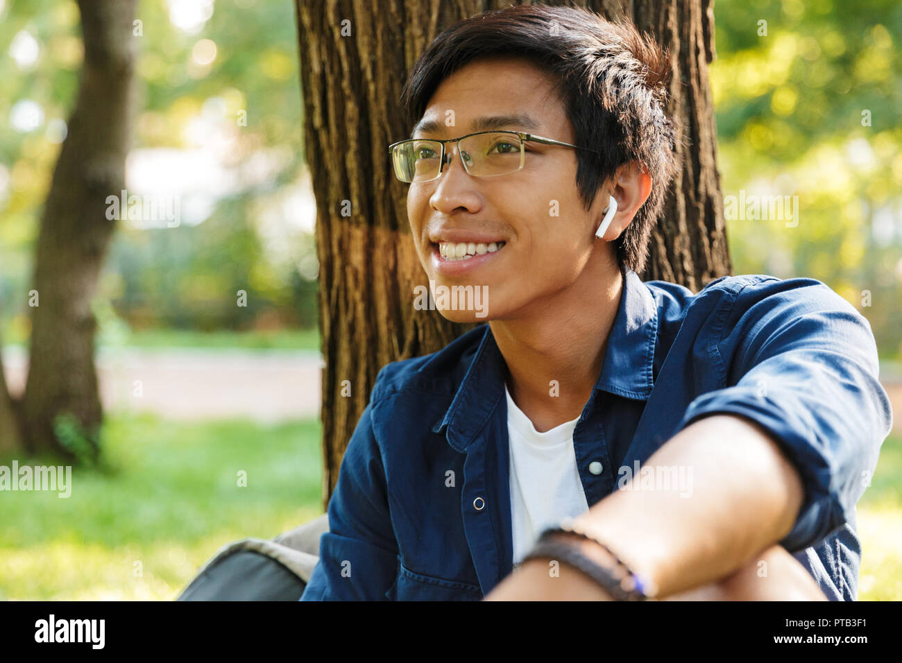 Happy asian male student in eyeglasses looking away while sitting near the  tree in park Stock Photo - Alamy