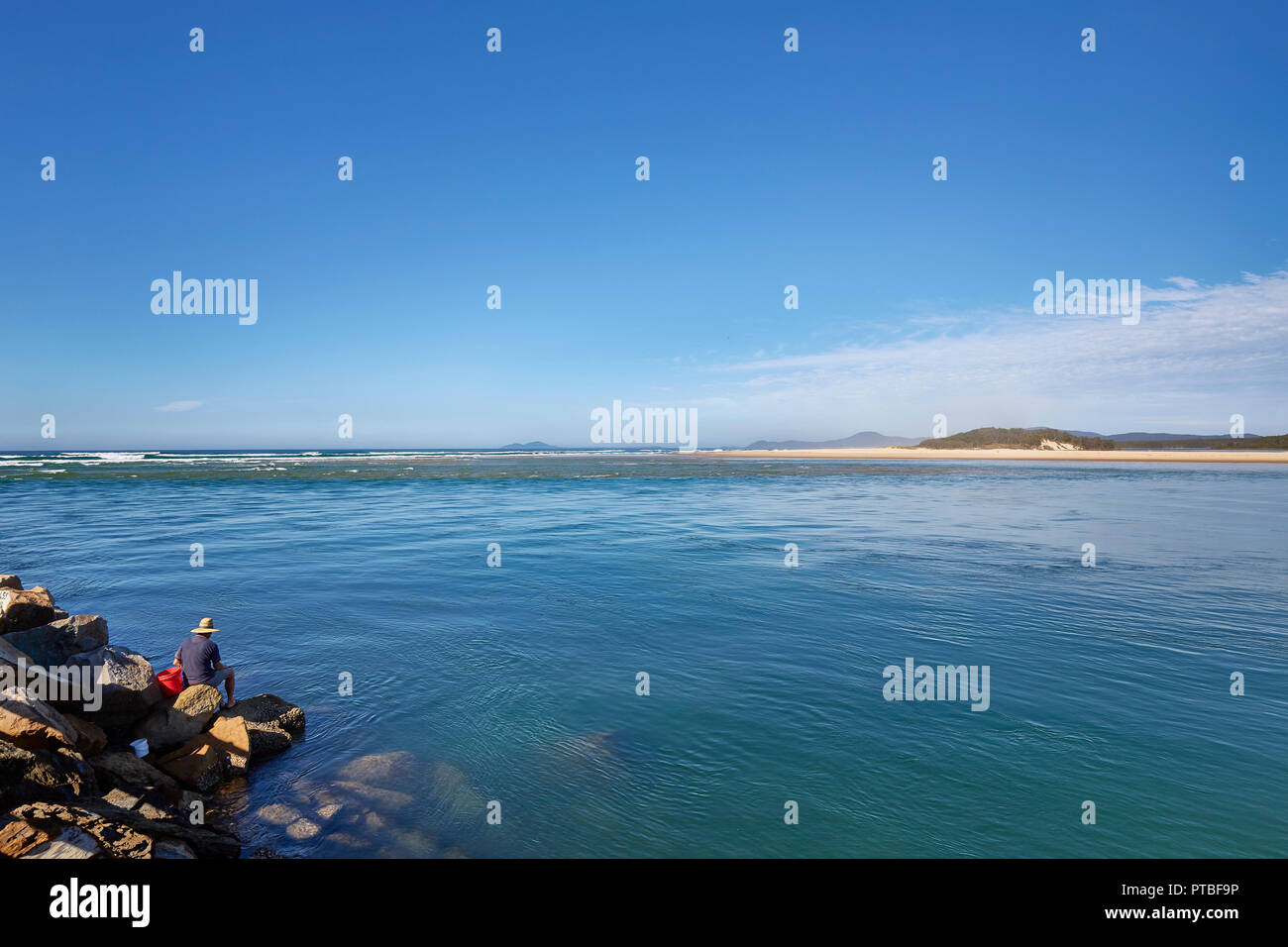 A man on his own wearing a hat sat on the boulders at the sea edge at Nambucca Heads, New South Wales, Australia Stock Photo