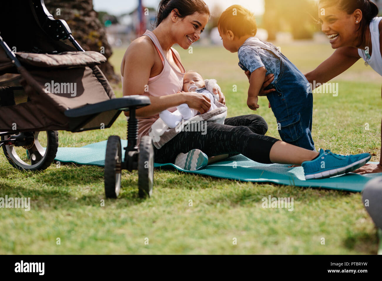 Smiling mothers sitting in park with their babies after the morning walk. Active moms with their kids spending time in park early in the morning. Stock Photo