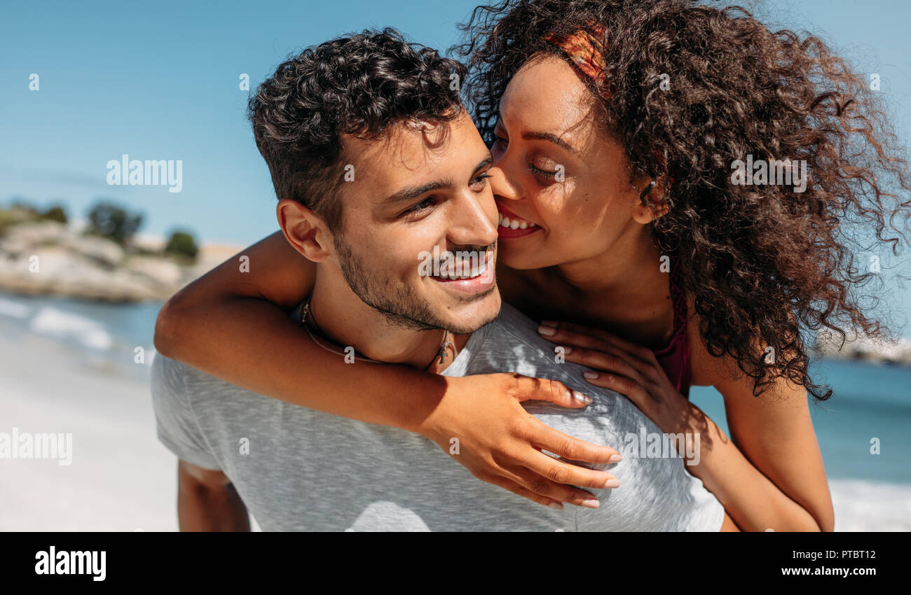 Smiling woman piggy riding on her boyfriend and kissing him. Man carrying his girlfriend on his back outdoors. Stock Photo