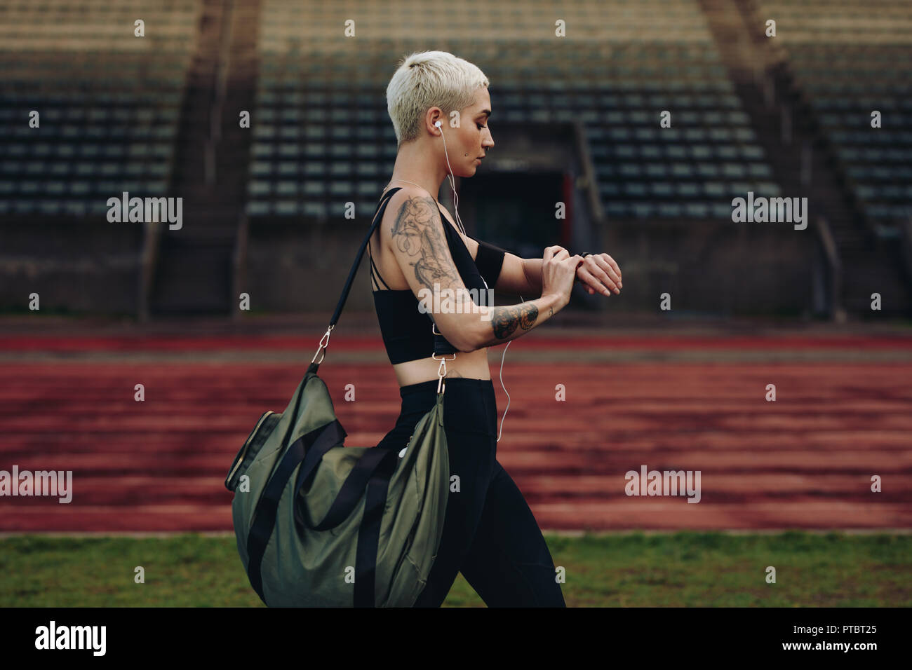 Female athlete walking beside an athletic track looking at her wrist watch. Woman in fitness wear walking in a track and field ground carrying her bag Stock Photo