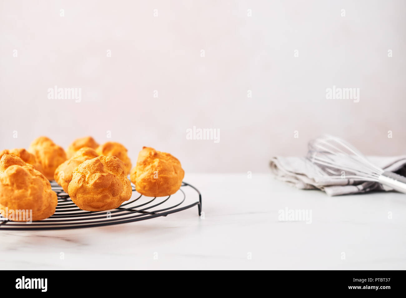 Bunch of fresh cream puff cakes on French wire rack with whisk and dishtowel on white marble table over pink background with copy space. Horizontal, s Stock Photo