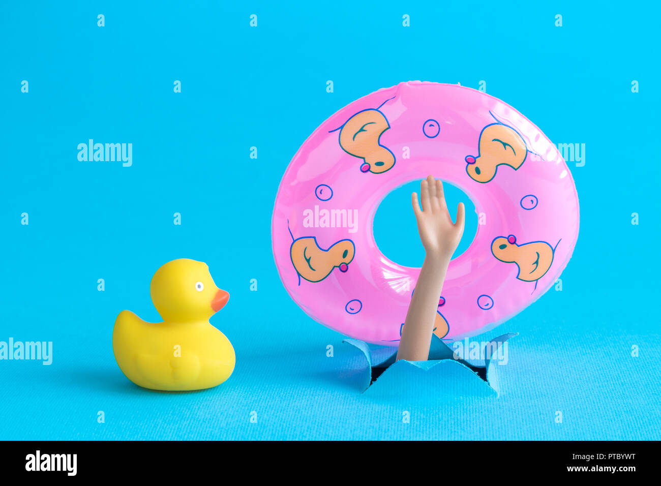 Rubber duck next to doll arm holding inflatable pool float and emerging from blue paper background. Drowning minimal creative abstract concept. Stock Photo
