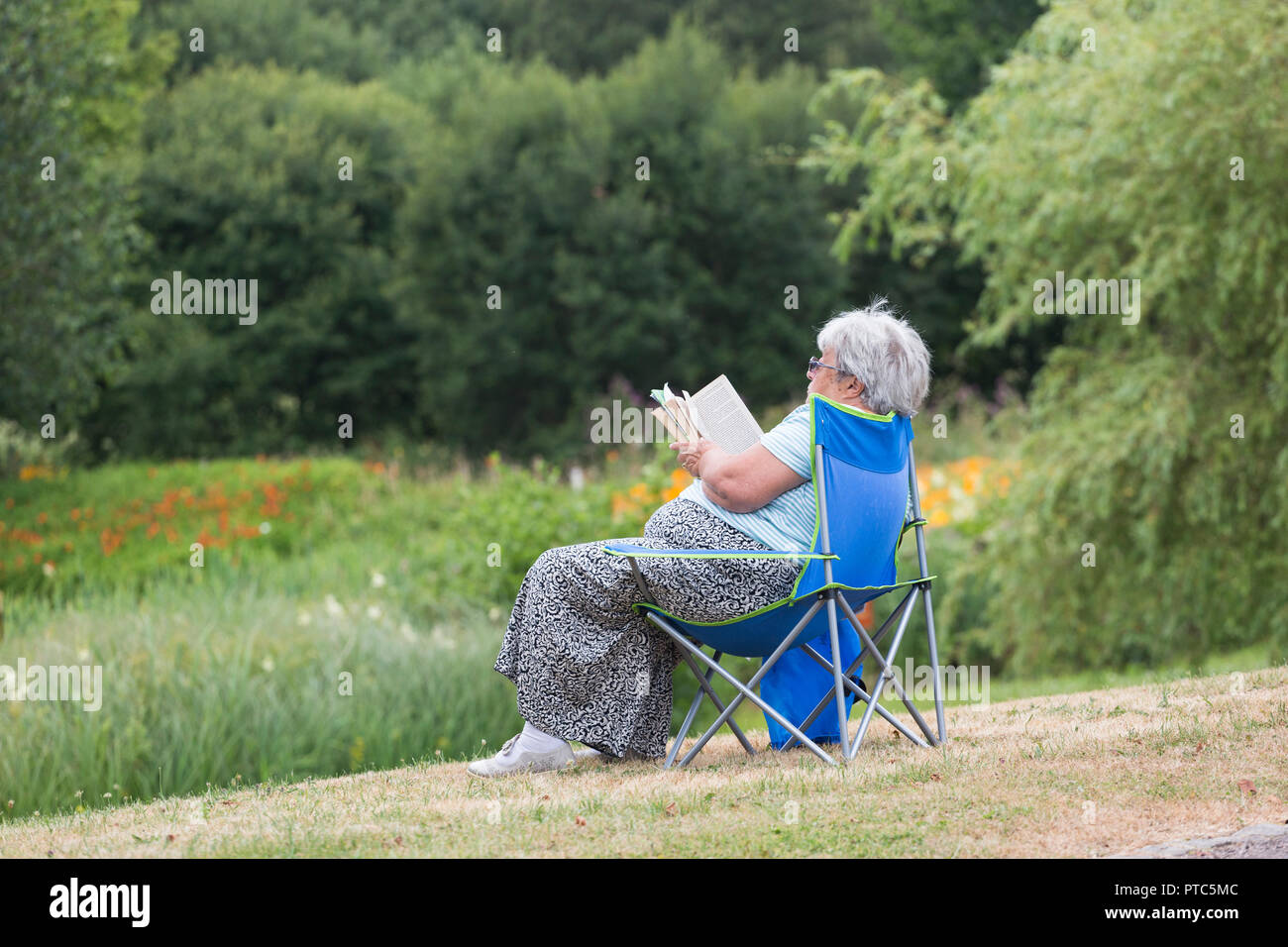 Woman sitting in a camping chair outdoors reads a book / novel. Stock Photo