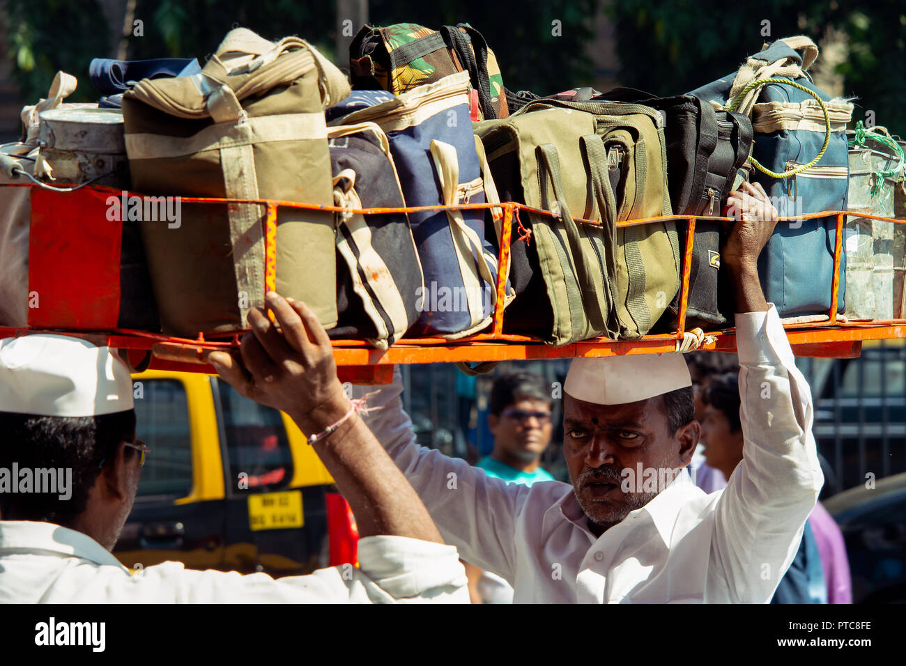 With the help of another a Indian man loads on a basket full of Dabbawala hot lunches on to his head to deliver around Mumbai, India. Stock Photo
