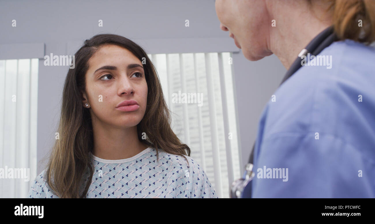 Mid aged nurse comforts young latina patient wearing hospital gown