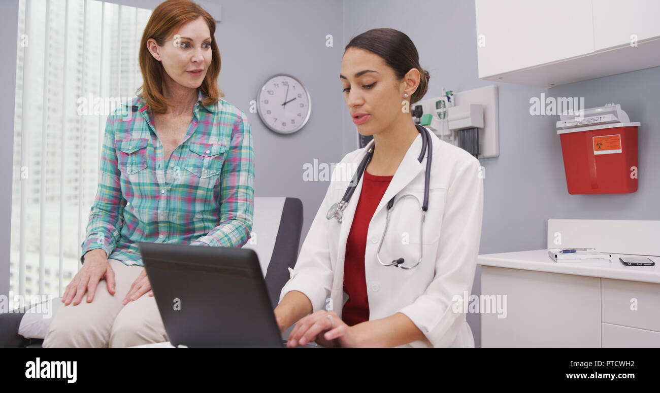 Young attractive latina doctor typing on laptop as she talks with senior patient Stock Photo