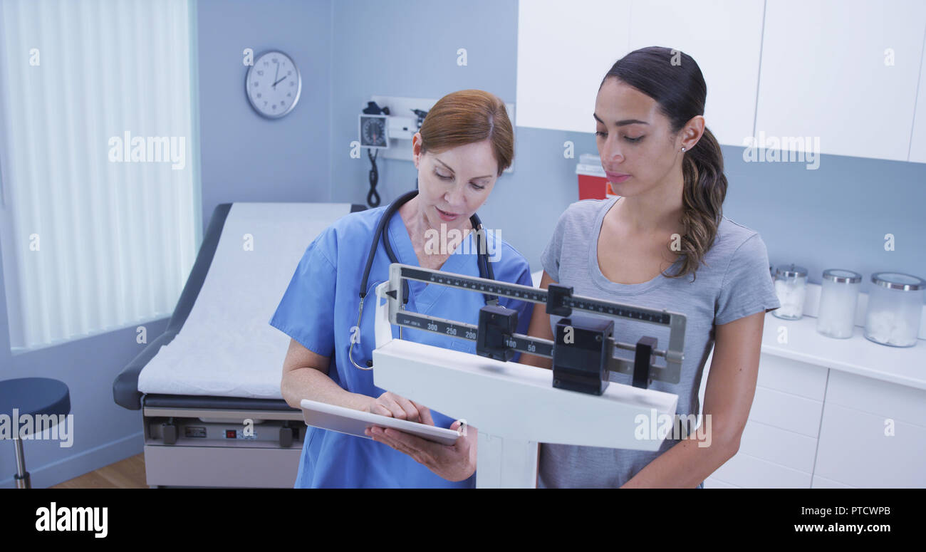 Charming middle aged nurse using scale to measure weight of young latina patient Stock Photo