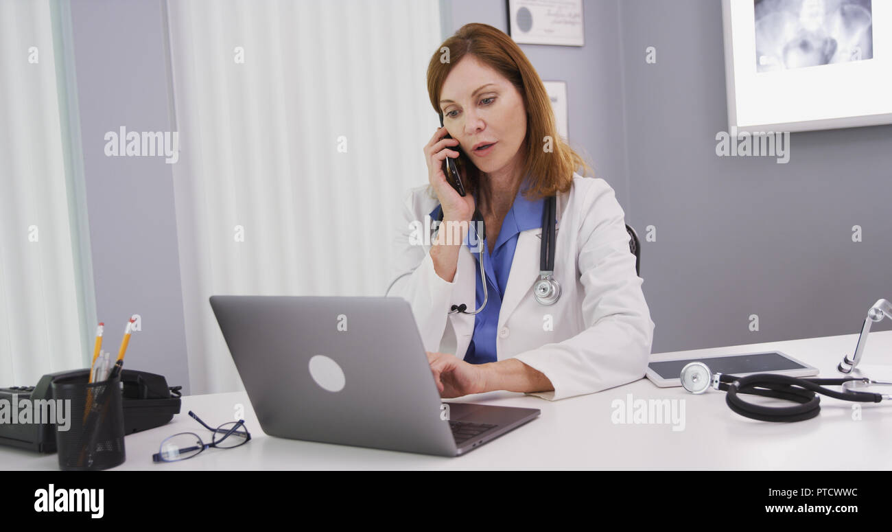 Attractive mid aged doctor using latop computer while talking on smartphone Stock Photo