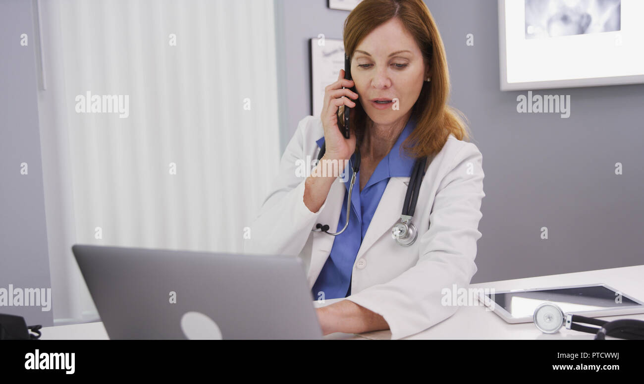Portrait of lovely female doctor using computer while she chats on phone Stock Photo