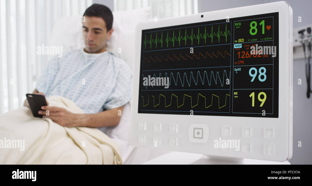 Close-up of EKG monitor with male patient using smartphone and lying down Stock Photo