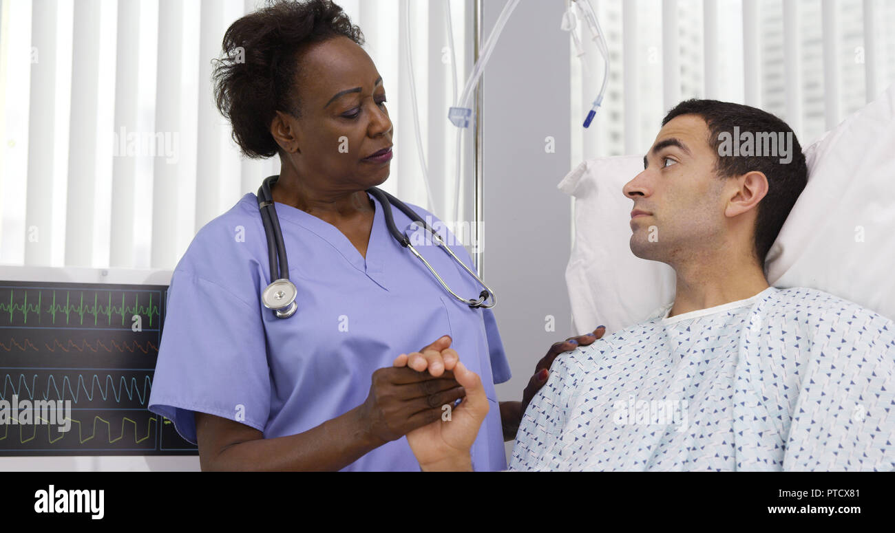 Senior black nurse comforting young hispanic patient on hospital bed Stock Photo