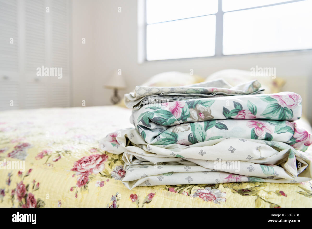 Stack of bedroom linen with floral, flower pattern on top of bed with quilt, quilted comforter, duvet, bright natural light from window, lamp, closet  Stock Photo