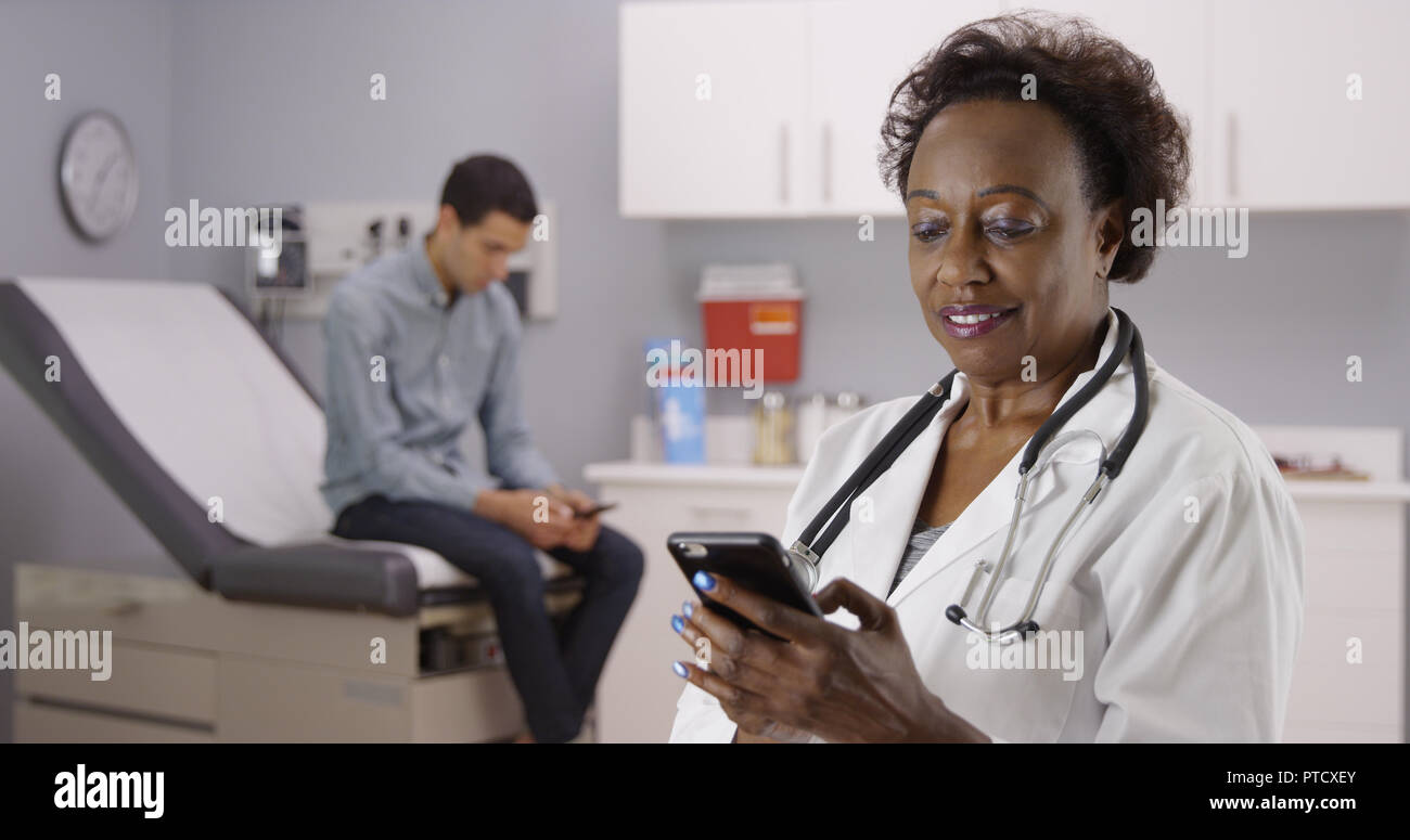 Middle aged African doctor using mobile phone to text while male patient waits Stock Photo