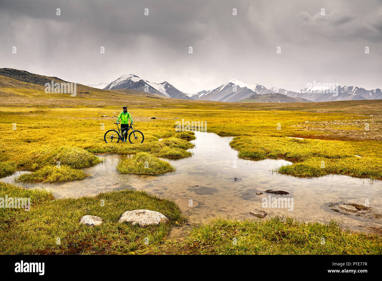 Man on mountain bike in green jacket near lake at stormy sky background. Stock Photo
