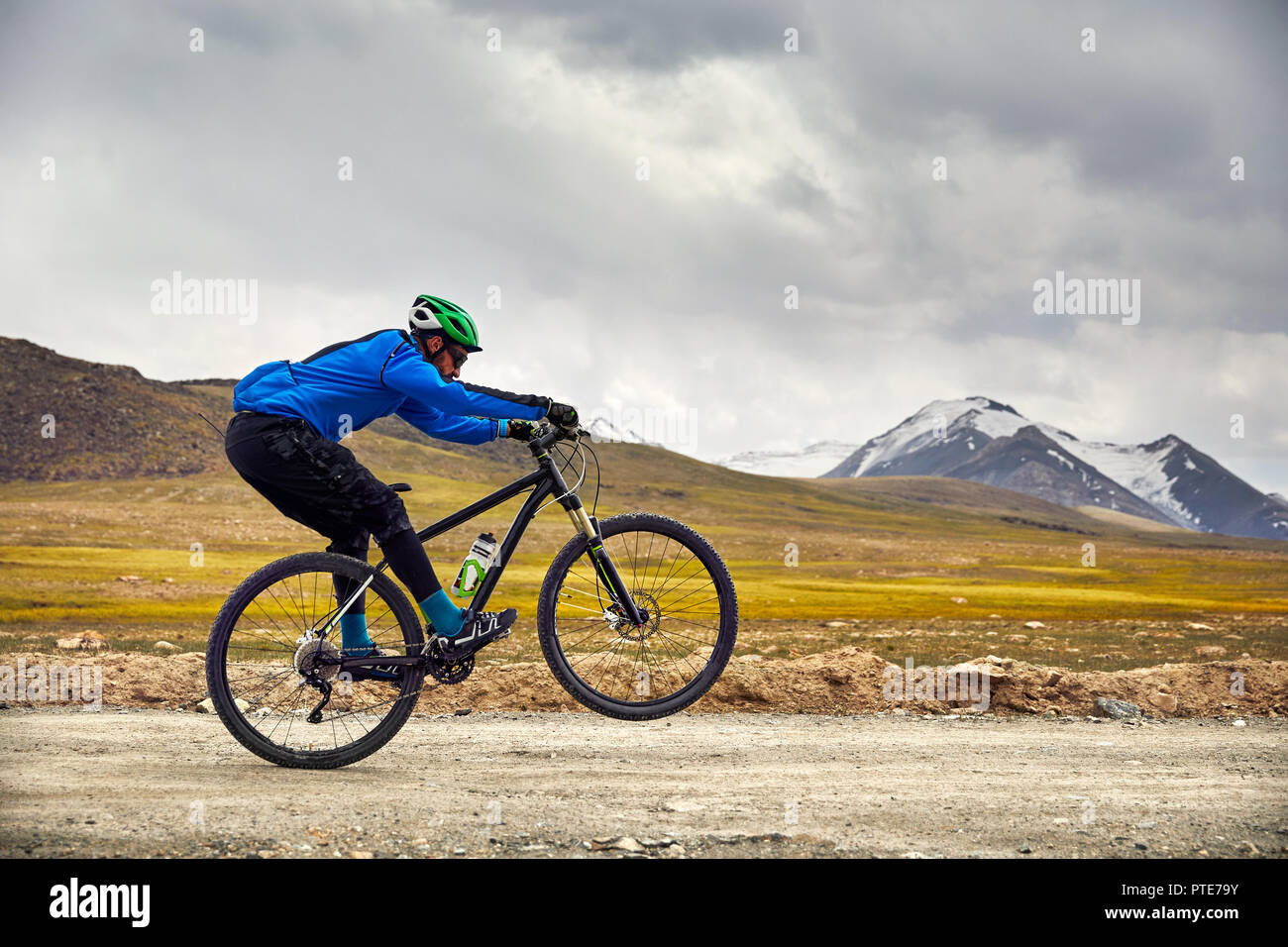 Man on mountain bike rides on the road in the high mountains against overcast sky background. Stock Photo