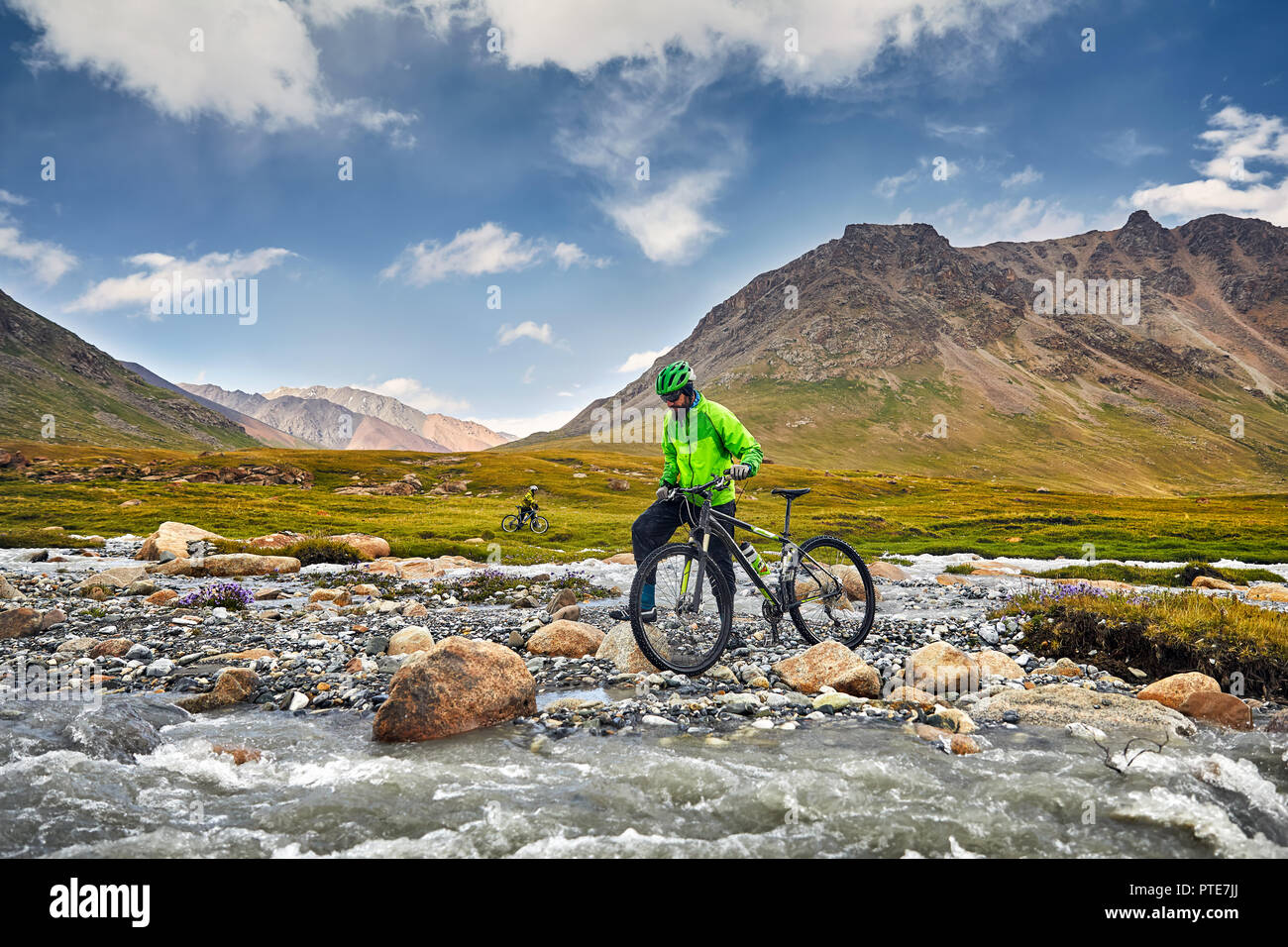 Man with mountain bike crossing the river in the wild mountains against cloudy sky background. Stock Photo