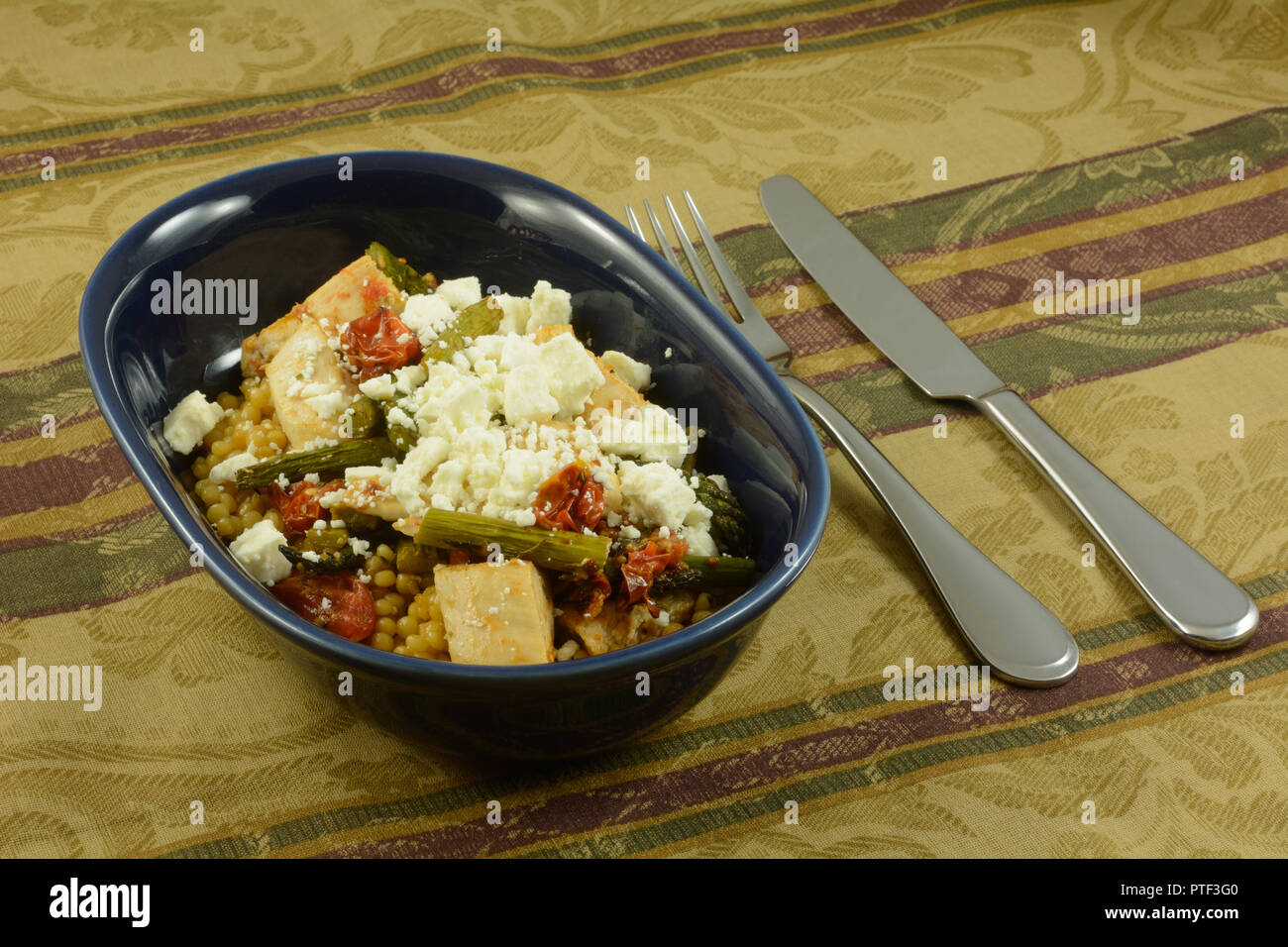 Israeli couscous or ptitim covered with chicken, roasted vegetables of asparagus and tomatoes with feta cheese on top in oval blue dish Stock Photo