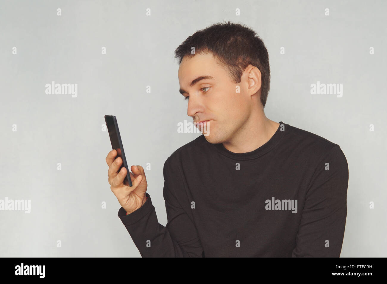 a young guy looks at the screen of a black smartphone with a dismissive emotion on his face. uninteresting correspondence. boring pastime. picture, ph Stock Photo
