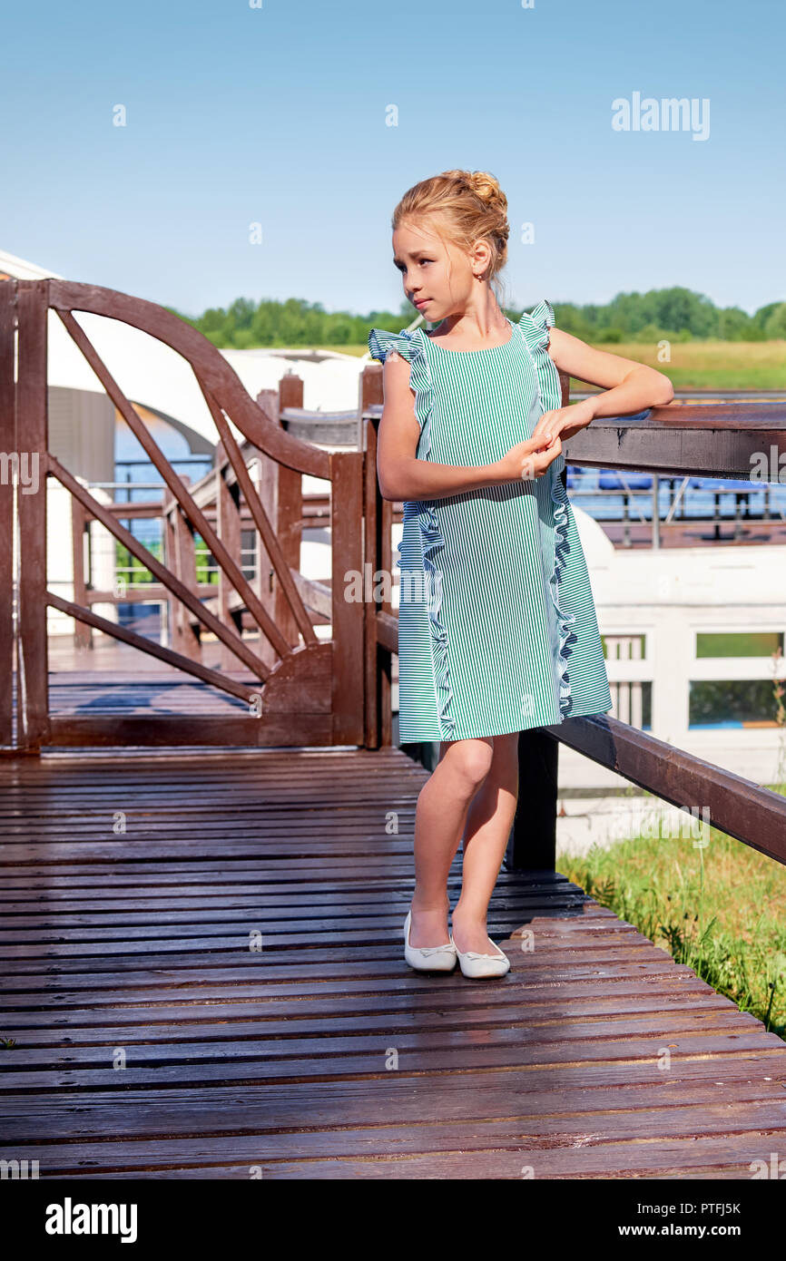 Fashionable summer attire, striped dress, marine style. Feminine pretty girl elegant turquoise dress for teenager standing leaning on bridge, looking away. Beautiful kid in cool clothes recreation. Stock Photo